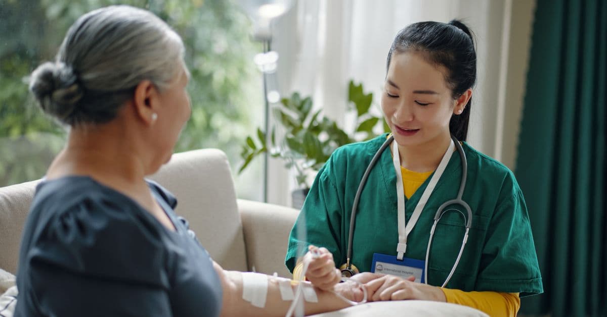 A nurse with dark hair in a ponytail and green scrubs administers an IV to an older woman as she sits on a couch.