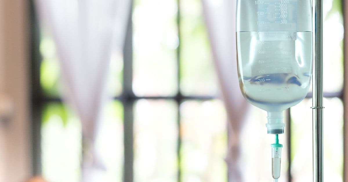 A close-up of an IV drip saline bag hanging in front of a residential window. The curtains are pulled back.