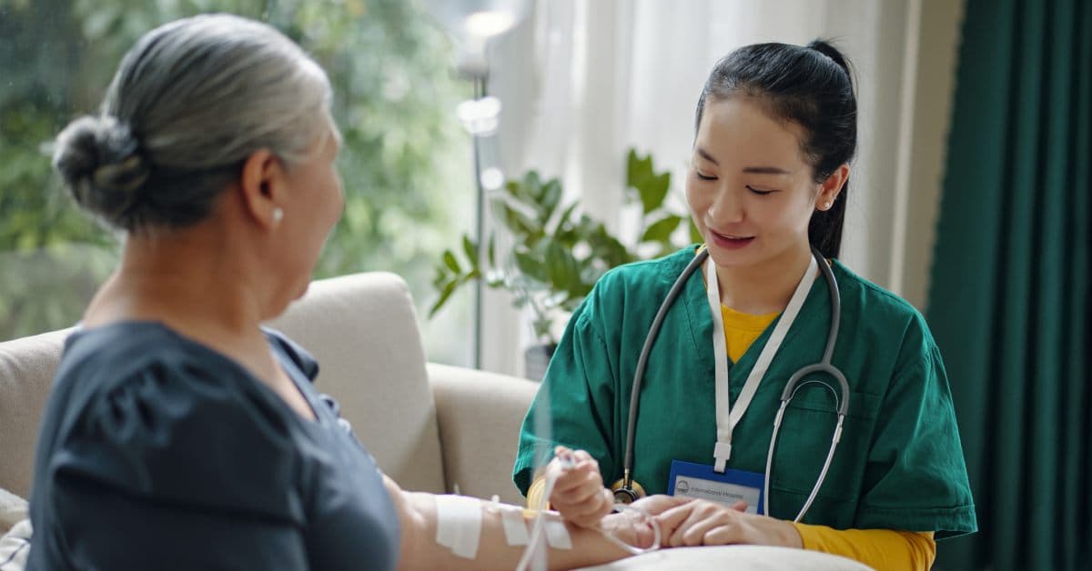 A registered nurse sits at the side of a senior woman's chair as she administers an IV drip through the woman's arm.