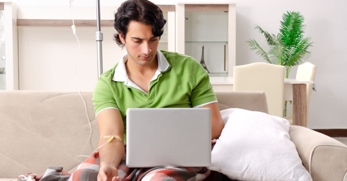 A young man works on a laptop and wears a green shirt as he receives a mobile IV drip in his home on the couch.