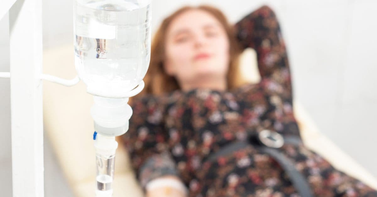 A close-up of a saline IV drip being adminstered to a female patient. The patient is behind the drip laying down.