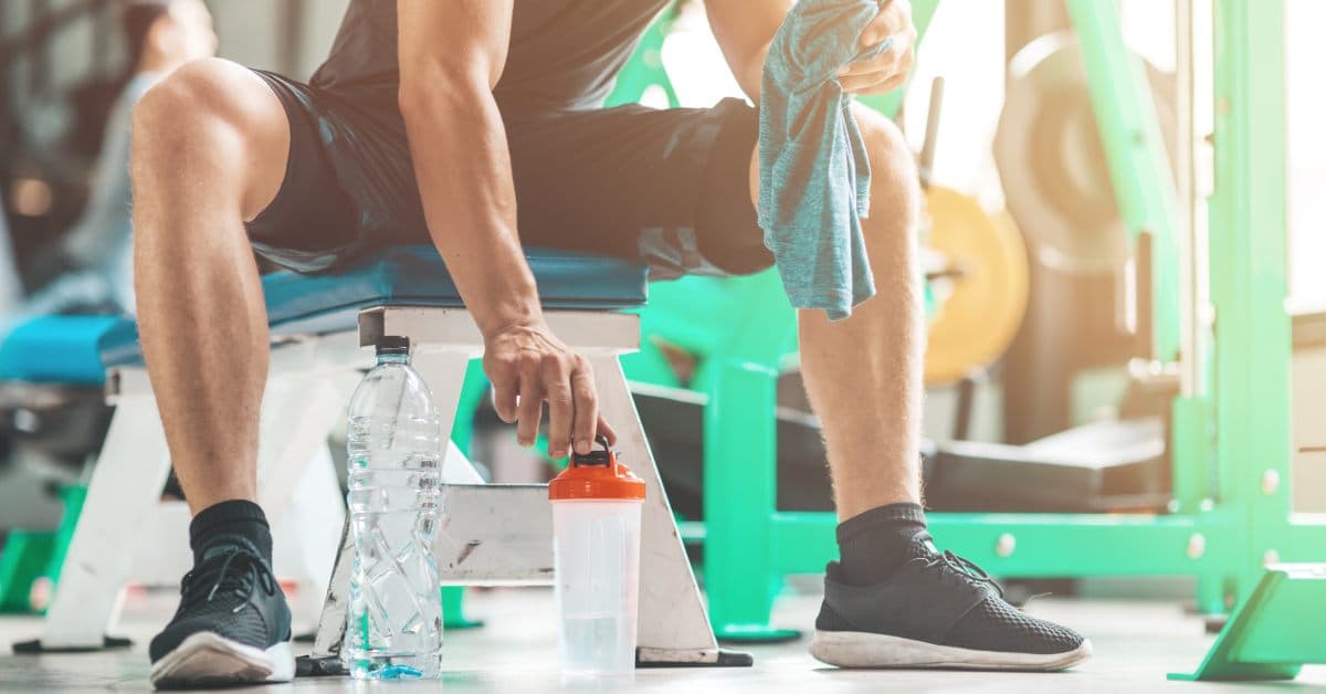 An athlete at the gym sits on a bench with a towel, water bottle, and shaker bottle on the floor in front of them.