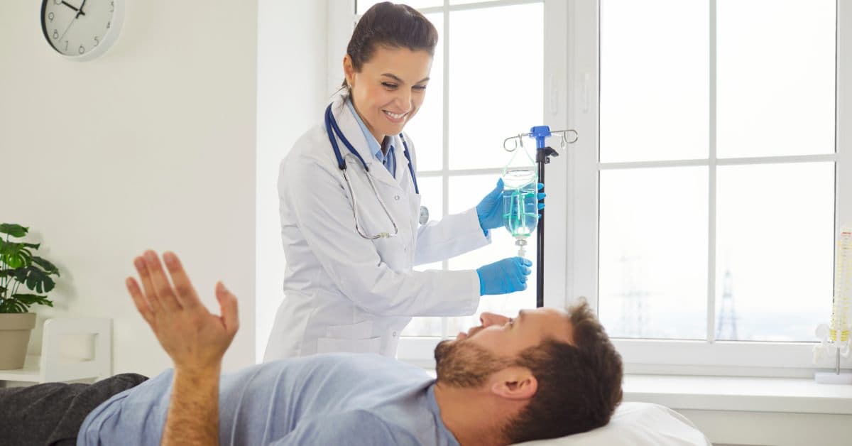 A nurse stands at the bedside of an athlete, getting ready to receive an IV drip therapy bag. The man talks with his hand.