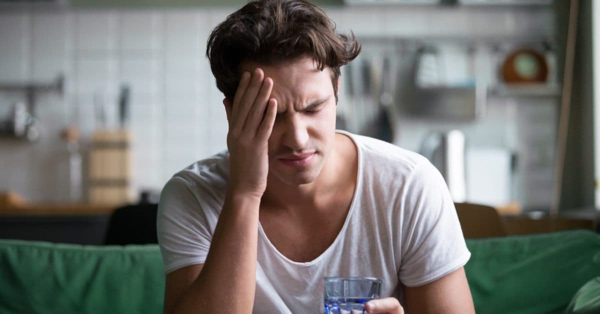 A young man in a white T-shirt sits on the edge of his couch as he holds his head with a glass of water in his hand.