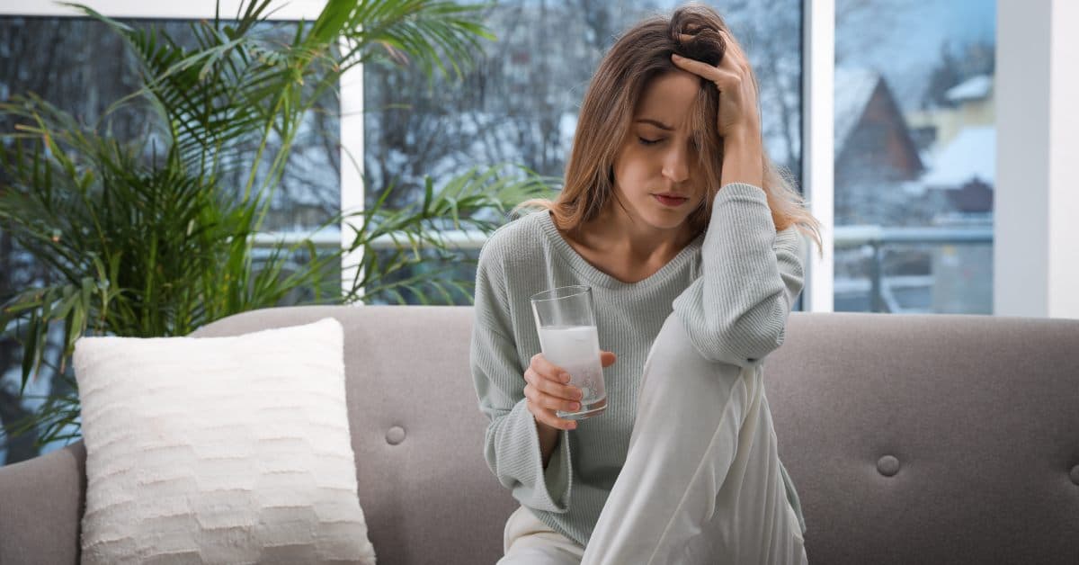 A young woman sits on her couch with a hangover, holding her head with her arm resting on her knee. She has a glass of water.