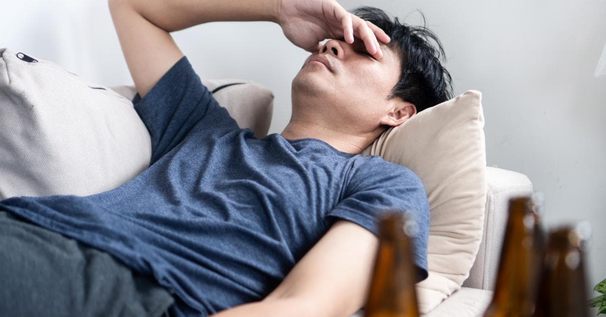 An exhausted man lays on the couch on his back with empty beer bottles on the coffee table across from him.