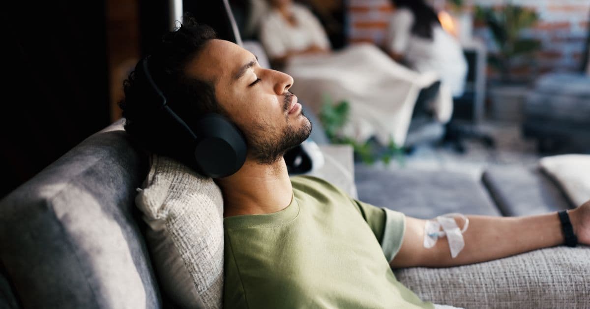 A man at home on the couch relaxing. He wears a headset and receives an IV therapy treatment in his arm.